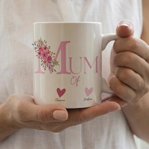 Peut inclure: Un mug en céramique blanc avec le texte rose Mum Of, orné d'un arrangement floral de fleurs roses et de feuilles vertes. Sous le texte se trouvent deux petits cœurs dans des tons de rose, avec les prénoms Florence et Melina inscrits dessous.