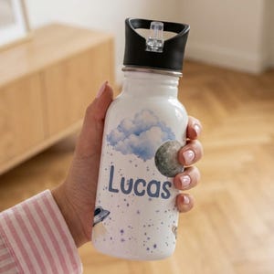 May include: A white water bottle featuring a space-themed design with a blue cloud, a gray moon, a small flying saucer, and scattered stars. The name Lucas is printed in blue letters on the front. The bottle has a black lid with a clear straw.