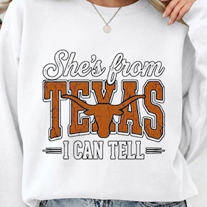 Puede incluir: Sudadera blanca con el texto "She's from TEXAS" en un diseño naranja y blanco desgastado. La palabra "TEXAS" presenta un gráfico de cuerno largo. Debajo del texto está "I CAN TELL" en blanco.
