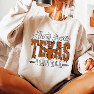 May include: Cream-colored sweatshirt with the text "She's from TEXAS - I CAN TELL" in brown and white. The design includes a longhorn graphic. The sweatshirt is a casual style.