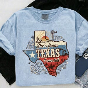 May include: Light blue t-shirt with a graphic of the state of Texas. The graphic includes a sunset, windmill, and the text "She's from Texas I can tell". The design is in red, white, and blue.
