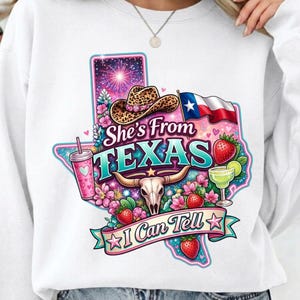Puede incluir: Sudadera blanca con un gráfico colorido que representa el estado de Texas. El diseño incluye un sombrero de vaquero, la bandera de Texas, fresas, una margarita y el texto "She's From TEXAS" y "I Can Tell".