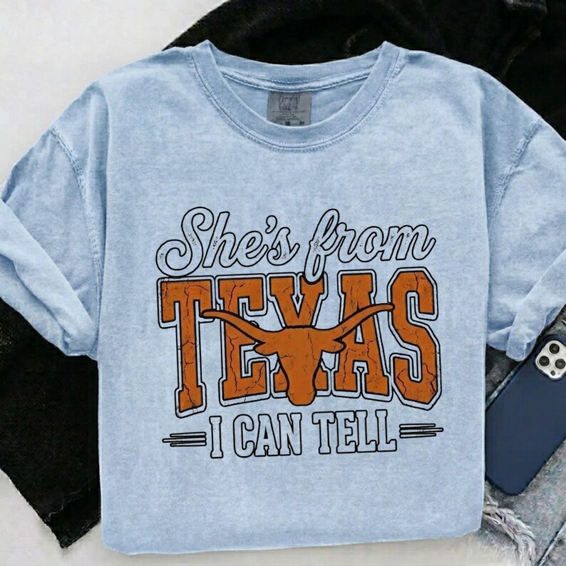 May include: Light blue t-shirt with the text "She's from TEXAS - I CAN TELL" in a distressed orange and white design. The shirt features a longhorn graphic, and is made of a soft, comfortable material.