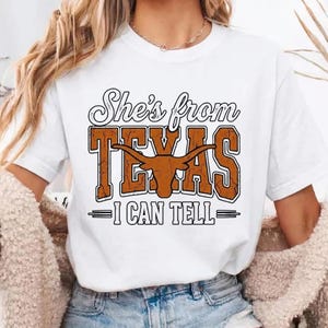 May include: White t-shirt featuring the text "She's from Texas - I CAN TELL" in a distressed orange and black design. The design includes a longhorn silhouette. The shirt is a casual, everyday wear item.