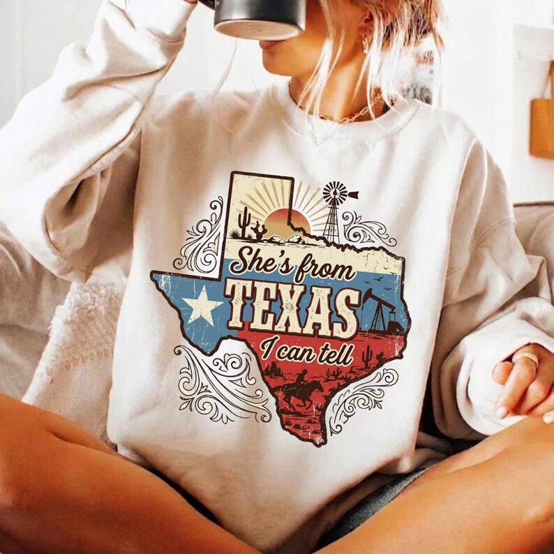 May include: Cream-colored sweatshirt with a graphic of the Texas state map. The map design includes a star, windmill, oil derricks, and a cowboy riding a horse. Text on the sweatshirt reads "She's from Texas I can tell."