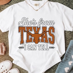 May include: White t-shirt with the text "She's from TEXAS - I CAN TELL" in brown and black. The design includes a longhorn silhouette. The shirt is a casual wear item.