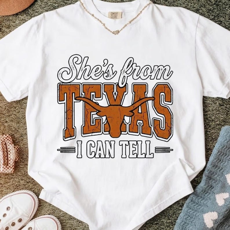 May include: White t-shirt with the text "She's from TEXAS - I CAN TELL" in brown and black. The design includes a longhorn silhouette. The shirt is a casual wear item.