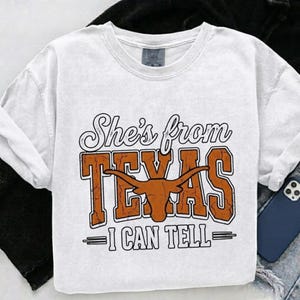May include: White t-shirt with the text "She's from TEXAS - I CAN TELL" in a distressed orange and white design. The design includes a longhorn silhouette. The shirt is a casual, unisex style.