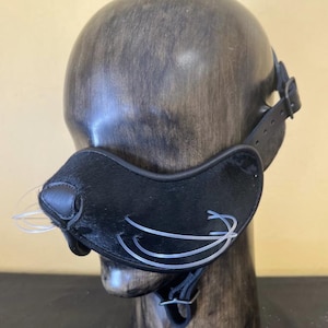 Puede incluir: Un bozal de cuero y piel negro con bigotes de plástico transparente se muestra en una forma de cabeza de madera. El bozal tiene una forma curva con una nariz y se asegura con correas de cuero.