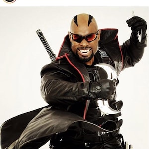 May include: A man in a black leather coat and sunglasses, holding two silver blades. He is smiling, with a sword on his back. The image is from the Instagram account eric_blade_brooks.