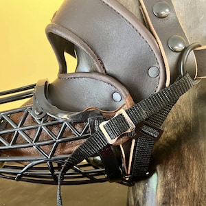May include: A brown leather and metal dog muzzle with a black wire cage front. The muzzle features adjustable straps and a buckle closure. The design includes a padded interior for comfort and is secured with rivets.