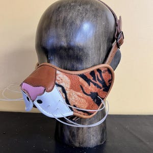 Puede incluir: Un bozal novedoso con un diseño de rayas de tigre, detalles de cuero marrón y una nariz rosa. El bozal se sujeta con una correa y hebilla de cuero marrón. Se exhibe en una forma de cabeza de madera.