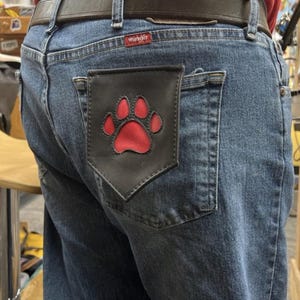 Puede incluir: Vaqueros azules con un bolsillo de cuero negro con un diseño de huella de pata roja. Los vaqueros tienen un cinturón de cuero marrón y la etiqueta de la marca Wrangler es visible.