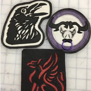 Könnte beinhalten: Drei Lederaufnäher mit verschiedenen Motiven: eine unregelmäßige Form mit einem schwarzen Raben auf weißem Grund, ein runder Aufnäher mit einem stierartigen Wesen in Violett und Weiß mit einem violetten Ring und ein quadratischer schwarzer Aufnäher.