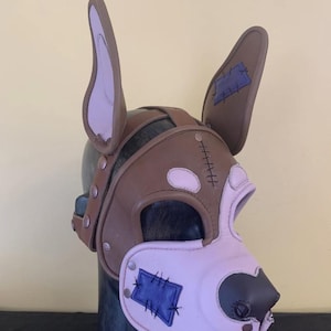 Puede incluir: Una m&aacute;scara de perro de cuero marr&oacute;n y rosa con detalles cosidos. La m&aacute;scara presenta orejas grandes, un hocico y parches azules. La m&aacute;scara se asegura con remaches y correas, y est&aacute; dise&ntilde;ada para adaptarse a la cabeza.