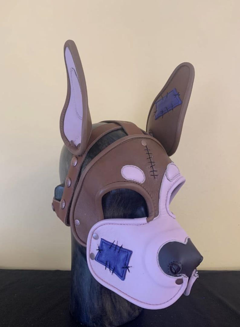 Puede incluir: Una m&aacute;scara de perro de cuero marr&oacute;n y rosa con detalles cosidos. La m&aacute;scara presenta orejas grandes, un hocico y parches azules. La m&aacute;scara se asegura con remaches y correas, y est&aacute; dise&ntilde;ada para adaptarse a la cabeza.