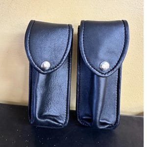 May include: Two black leather pouches with rounded flaps and silver snap closures. The pouches are rectangular with a curved top and are likely designed to hold small items. The background is a light tan color.