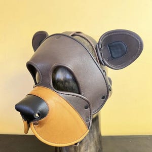 Könnte beinhalten: Eine braune Lederbärenmaske mit schwarzer Nase und hellbrauner Schnauze. Die Maske hat angebrachte Ohren und ist mit Nieten und Riemen befestigt. Die Maske ist für den Kopf konzipiert.