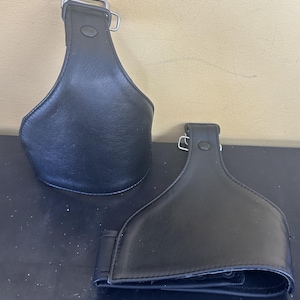 Peut inclure: Deux couvre-étriers en similicuir noir en forme de goutte, dotés d'une boucle en métal argenté sur le dessus et d'un bouton-pression noir. Les articles présentent des bords cousus et une finition mate lisse sur la partie extérieure.