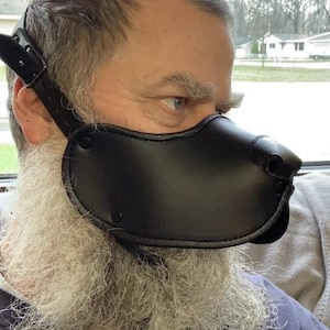Könnte beinhalten: Schwarze Leder-Hundemaulkorb mit verstellbaren Riemen und Nietendetails. Der Maulkorb bedeckt den Nasen- und Mundbereich und wird mit einem Riemen um den Kopf befestigt. Das Produkt ist so gestaltet, dass es einer Hundenase ähnelt.
