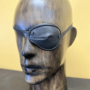 May include: A black leather eye patch with an elastic strap, displayed on a wooden mannequin head. The eye patch is oval-shaped and covers the left eye. The mannequin has a dark, weathered finish.
