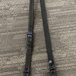 May include: Black leather strap with an adjustable buckle and rivets. The strap has a rectangular shape and is designed for use with a holster or other equipment. The strap is made of a durable material.