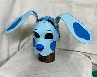 May include: A light blue and dark blue dog-shaped hood featuring long, floppy ears with dark blue accents, a dark blue nose, and cutouts for the eyes and mouth. The hood is constructed with stitched panels and metal rivets, secured by straps.