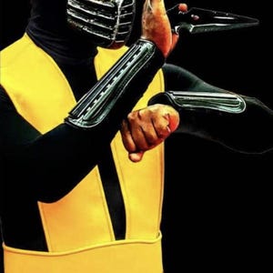 May include: A person in a black and yellow costume, with a black hood and a silver mask. The costume includes black sleeves with silver accents and a yellow sash. The person is holding a black weapon, with a black background.