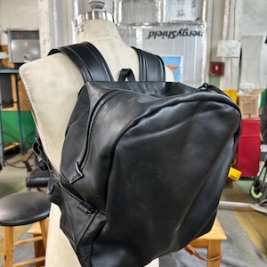 Peut inclure: Un sac à dos en cuir noir avec un compartiment principal zippé et des bretelles réglables. Le sac à dos a une poche avant rectangulaire et une poignée supérieure. Le sac est présenté sur un mannequin.