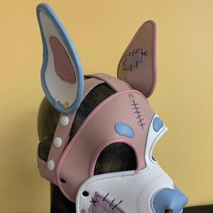 Puede incluir: Una m&aacute;scara de perro de cuero rosa, blanco y azul con detalles cosidos. La m&aacute;scara presenta orejas grandes, un hocico y una correa con broches blancos. El dise&ntilde;o incluye parches y acentos azules.