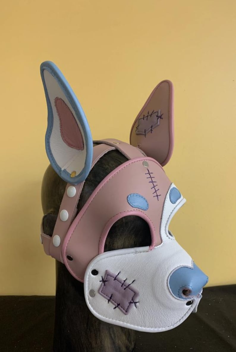 Puede incluir: Una m&aacute;scara de perro de cuero rosa, blanco y azul con detalles cosidos. La m&aacute;scara presenta orejas grandes, un hocico y una correa con broches blancos. El dise&ntilde;o incluye parches y acentos azules.