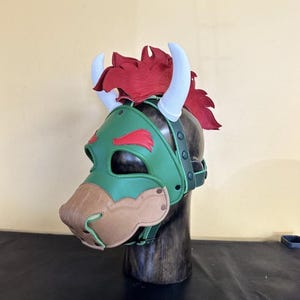 Könnte beinhalten: Eine grüne und braune Bullenmaske mit weißen Hörnern und rotem Haar. Die Maske ist aus Leder gefertigt und hat ein detailliertes Design, einschließlich einer Schnauze und Augenausschnitten. Die Maske wird auf einem Holzständer ausgestellt.