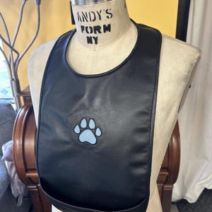 May include: Black bib with a rounded neckline and a pocket at the bottom. A light blue paw print is embroidered on the front. The bib is displayed on a mannequin with the text "ANDY'S FORM NY".