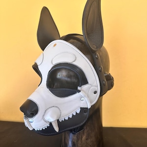 May include: A black and white leather dog mask with large, upright ears. The mask features a white muzzle with black accents, including a nose and eye openings. The design includes a row of teeth and is secured with straps and rivets.
