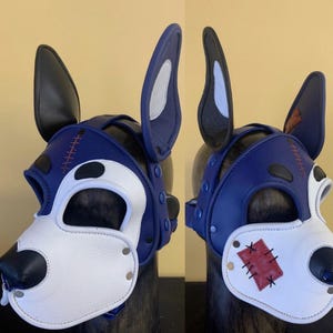Puede incluir: Una m&aacute;scara de perro de cuero azul y blanco con orejas negras. La m&aacute;scara presenta un hocico blanco, una nariz negra y detalles de costura rojos. Las orejas est&aacute;n forradas con tela blanca y gris. La m&aacute;scara se asegura con correas y remaches.