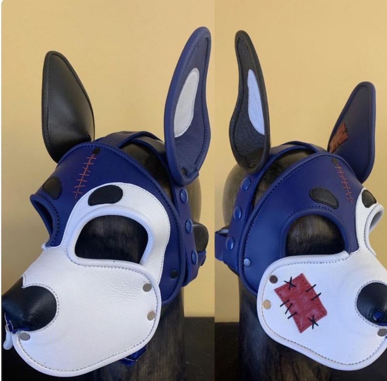 Puede incluir: Una m&aacute;scara de perro de cuero azul y blanco con orejas negras. La m&aacute;scara presenta un hocico blanco, una nariz negra y detalles de costura rojos. Las orejas est&aacute;n forradas con tela blanca y gris. La m&aacute;scara se asegura con correas y remaches.