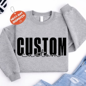 May include: A heather grey crewneck sweatshirt featuring the word CUSTOM in a large, bold, black sans-serif font, with the word custom written underneath in a smaller, black, cursive script font. The garment has long sleeves and ribbed cuffs.