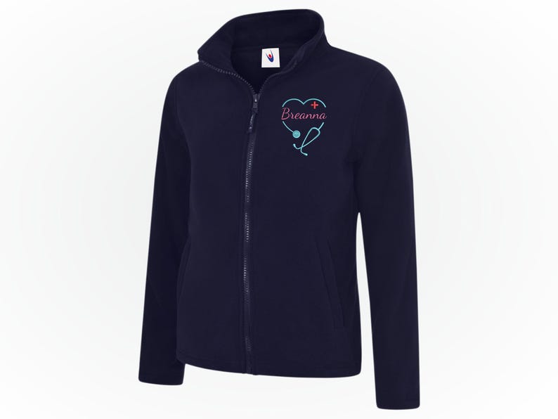 May include: Navy blue fleece jacket with a full zipper and a high collar. The jacket features a heart-shaped design with a stethoscope and the name "Breanna" embroidered in pink and teal. A comfortable and warm outerwear option.