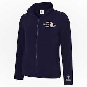 Könnte beinhalten: Marineblaue Fleecejacke mit durchgehendem Reißverschluss und hohem Kragen. Auf der Brust ist der Text "THE NURSE FACE" mit einer Stethoskop-Grafik und auf dem Ärmel der Name "Sutton" abgebildet.