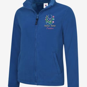 May include: Royal blue fleece jacket with a full zipper and a high collar. The jacket features an embroidered stethoscope and floral design with the text "Senior Sister Preston". A pocket is visible on the lower left side.
