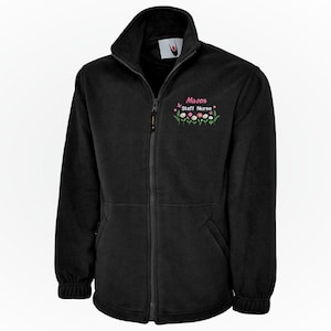 Könnte beinhalten: Schwarze Fleecejacke mit durchgehendem Reißverschluss, hohem Kragen und gesticktem Blumendesign. Der Text "Mason Staff Nurse" ist über dem Blumendesign gestickt. Die Jacke hat zwei Vordertaschen und gerippte Bündchen.