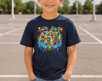 May include: A navy blue short-sleeve t-shirt featuring a colourful graffiti-style graphic with the text I WAS NOW I'M 67. The design includes bright paint splatters, a yellow lightning bolt, a smiley face, and two blue hands holding the number 67.