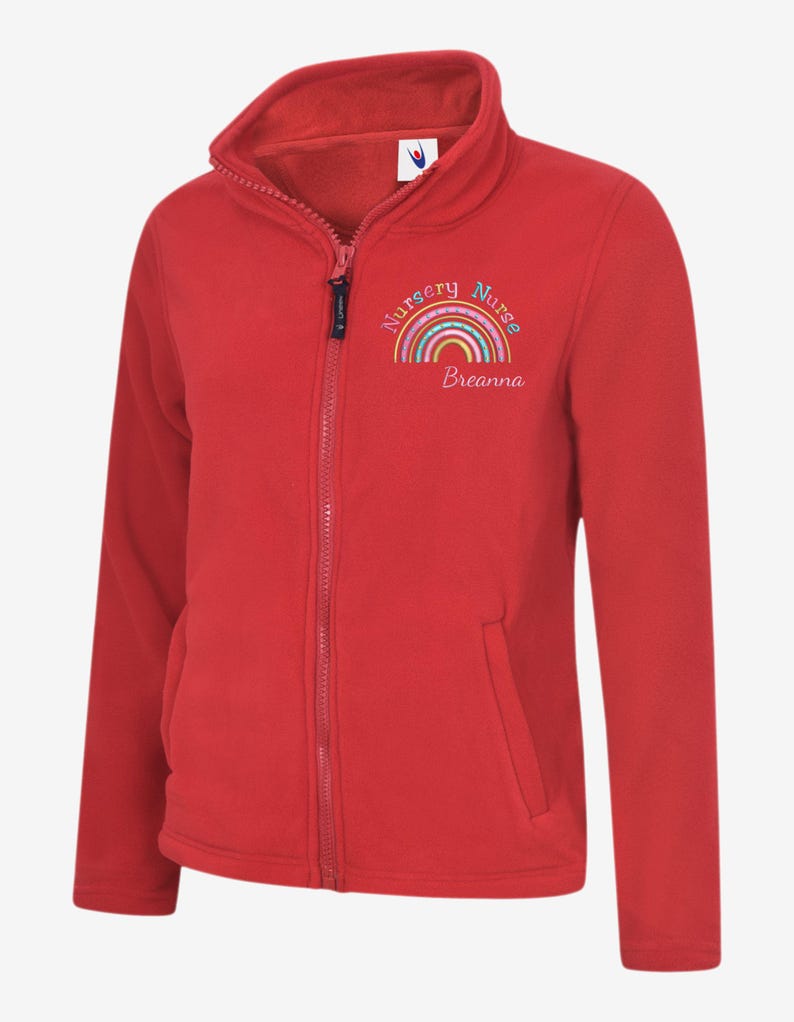 May include: A red fleece jacket with a full zipper and a high collar. The jacket features the text "Nursery Nurse" above a rainbow design, with the name "Breanna" below the rainbow. The jacket has two front pockets.