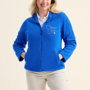 May include: A vibrant blue fleece jacket with a full zipper and two front pockets. The jacket features the text "Nurse Jackson" with a stethoscope graphic in pink. The model is wearing khaki pants.