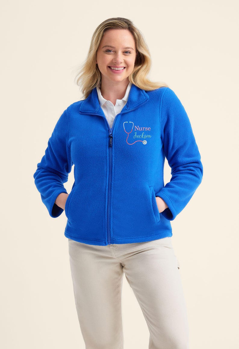 May include: A vibrant blue fleece jacket with a full zipper and two front pockets. The jacket features the text "Nurse Jackson" with a stethoscope graphic in pink. The model is wearing khaki pants.