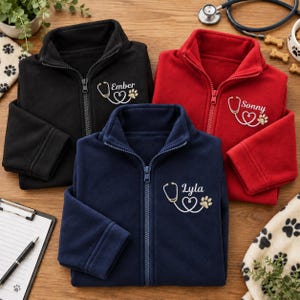 Pode incluir: Três casacos de fleece com fecho em preto, vermelho e azul-marinho. Cada casaco apresenta um bordado no peito esquerdo composto por um estetoscópio em forma de coração, uma pegada de pata e um nome: Ember, Sonny ou Lyla.