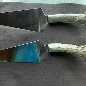 May include: Two stainless steel cake servers with antler handles. The blades are wide and reflective, and the handles are a light beige color with a textured, naturalistic design. The servers are displayed on a dark brown surface.