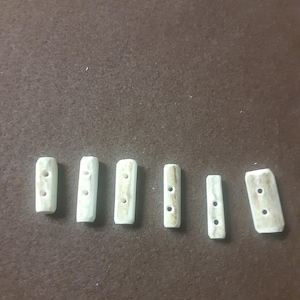 May include: A collection of six rectangular, off-white antler buttons with two or three holes each. The buttons are arranged on a brown surface, showcasing their natural color variations and textures. These buttons are ideal for crafting and sewing projects.