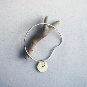May include: A silver metal bracelet with a circular design and a small, round, gold-coloured pendant. The bracelet is displayed on a piece of driftwood against a light grey background. The bracelet's diameter is approximately 6.35 cm.