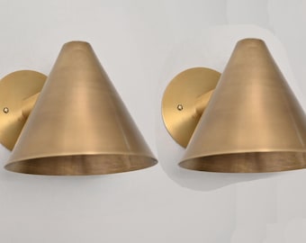 May include: Two brass wall sconces with a conical shade design. Each sconce features a circular backplate and a cone-shaped shade. The sconces are made of a warm, metallic brass material and are mounted on a white wall.